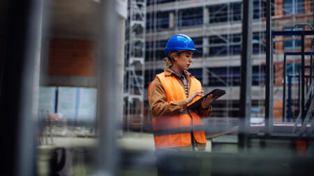 A woman in an orange vest and hard hat stands in front of a building, overseeing construction progress.