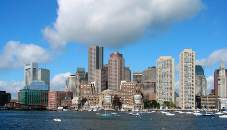 A panoramic view of Boston's skyline featuring high-rise buildings, with a waterfront marina under a blue sky with scattered clouds.