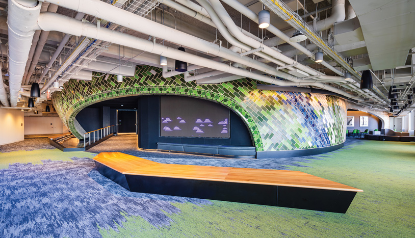 A colorful section within Google's headquarters featuring colorful carpet, a place to sit, and a large projector screen.