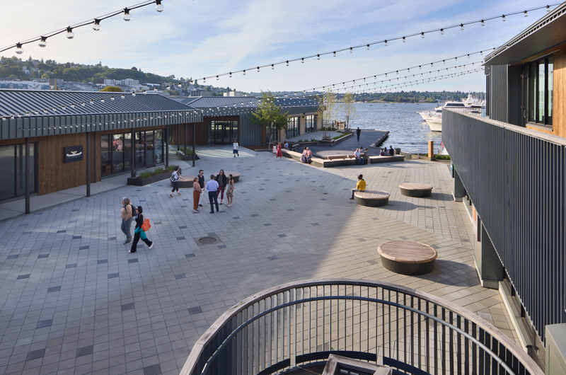 People stroll along a waterfront area, with a prominent large building in the background, creating a vibrant urban scene.