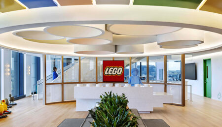 LEGO US HQ image of Logo, reception desk, Lego character and wall of windows with grey bench area center and potted plants built by Structure Tone Boston