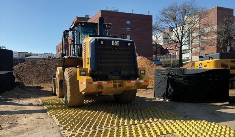 A large yellow bulldozer on a construction site, parked on a textured yellow ground mat. Piles of dirt, buildings, and a tree are in the background.