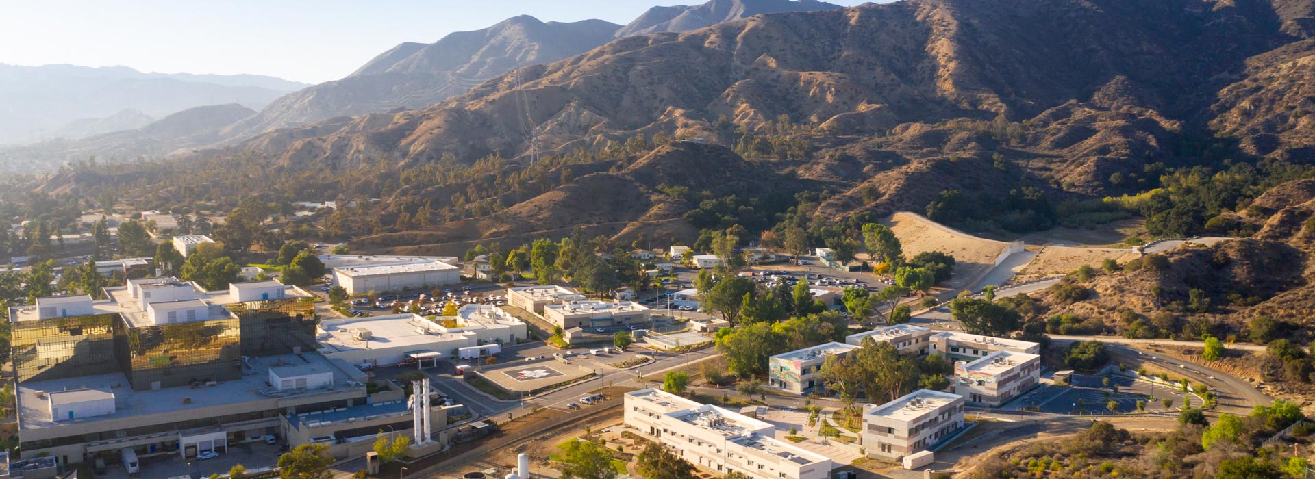 Aerial view of several low-rise buildings with light-colored roofs are connected by roads, parking areas, and landscaped green spaces.