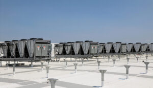 Working Through Complexity in Equipment Procurement A row of large industrial HVAC cooling units sits on a bright white rooftop, supported by a grid of metal posts and walkways. The equipment stretches across the frame beneath a clear blue sky, creating a clean, geometric pattern with strong horizontal lines and an expansive, open-air setting.