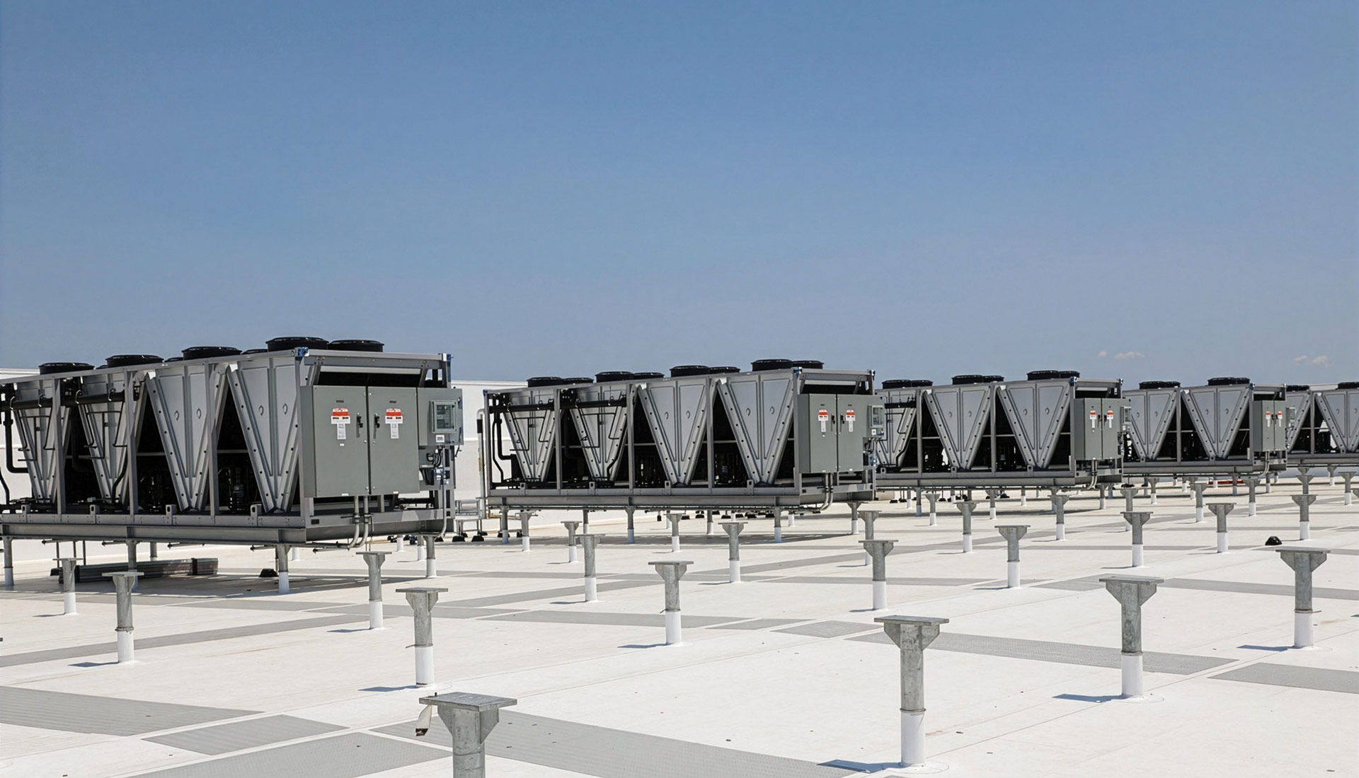 A row of large industrial HVAC cooling units sits on a bright white rooftop, supported by a grid of metal posts and walkways. The equipment stretches across the frame beneath a clear blue sky, creating a clean, geometric pattern with strong horizontal lines and an expansive, open-air setting.