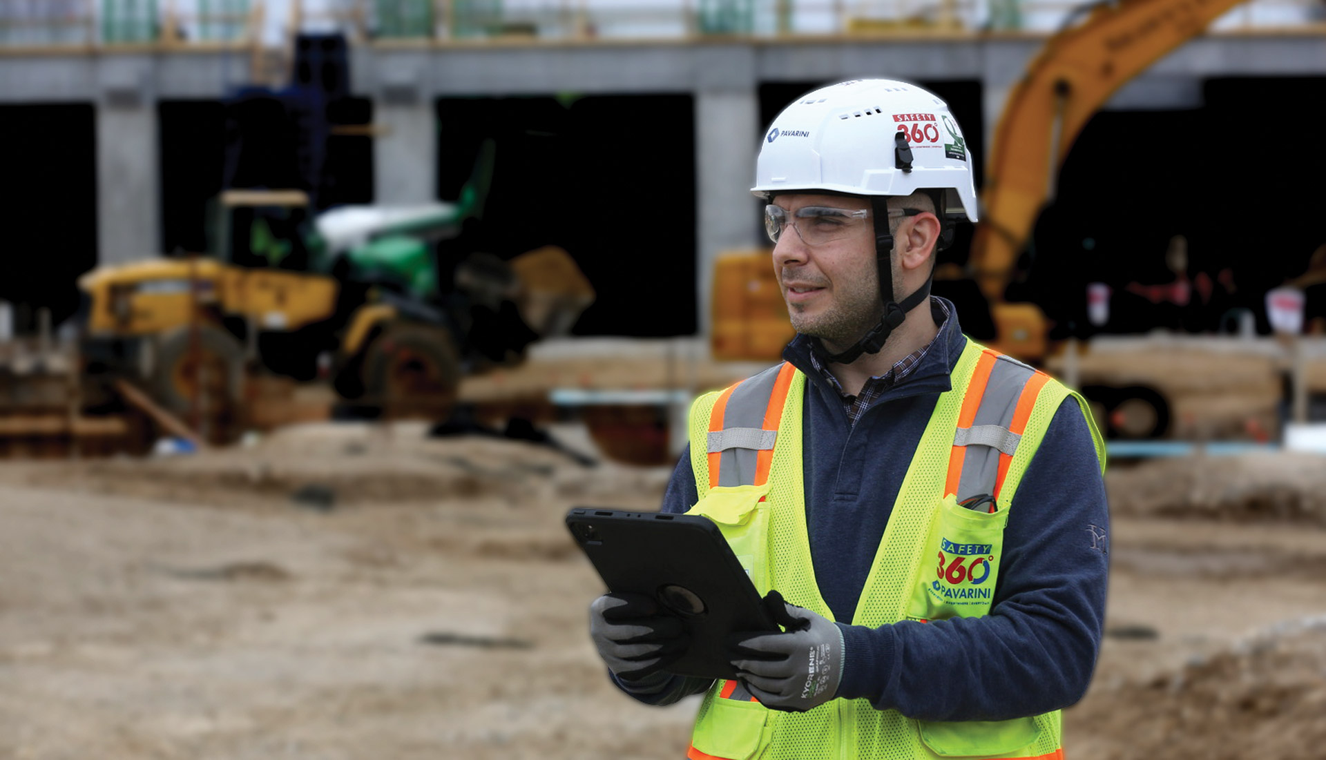 A construction site supervisor wearing a white hard hat, safety glasses, gloves, and a high-visibility reflective vest stands in the foreground holding a tablet. Heavy construction equipment and a partially built structure are visible out of focus in the background, indicating an active construction site.