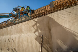 Integrating Climate Risk into Construction: From Assessment to Action Construction workers wearing hard hats and high-visibility safety vests stand in a blue aerial lift platform near the top edge of a large concrete wall.