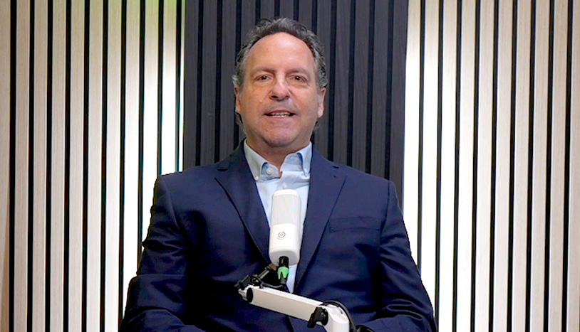 Pete Kobelt in a navy suit sits behind a white microphone in a recording studio with black-and-white acoustic wall panels, speaking directly to the camera.