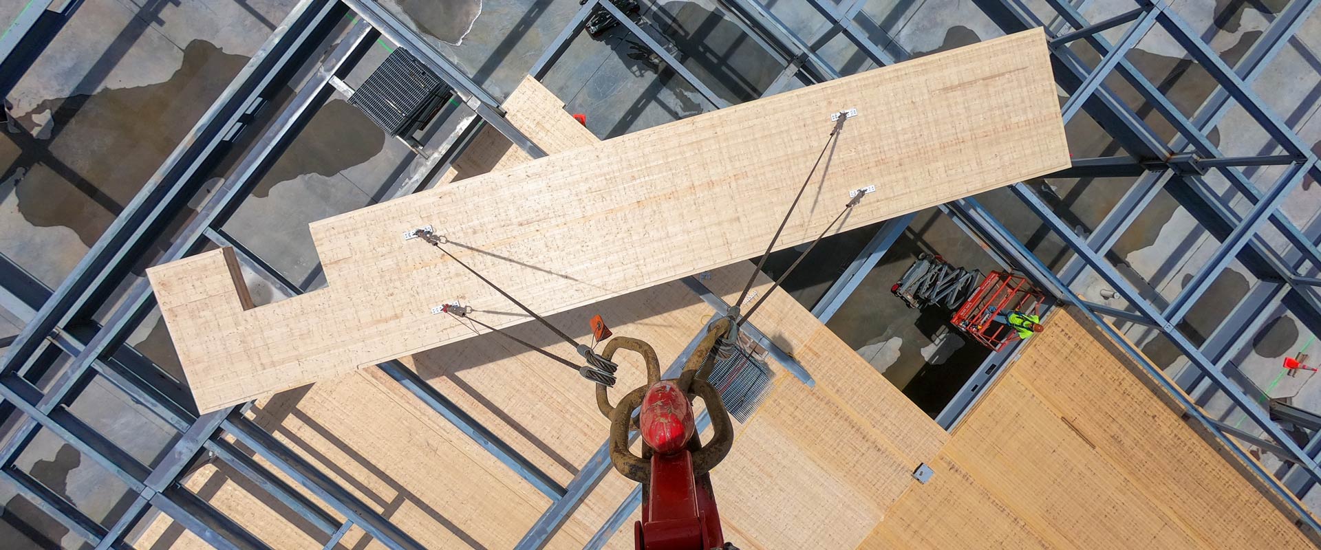 A crane lifts a large mass timber panel into place within a steel building frame as workers guide the installation from below, viewed from directly overhead.