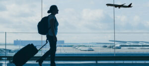 The Future of Airport Construction: Three Trends Shaping What’s Next Silhouetted traveler with a backpack pulling a suitcase through an airport terminal as a passenger plane approaches outside.