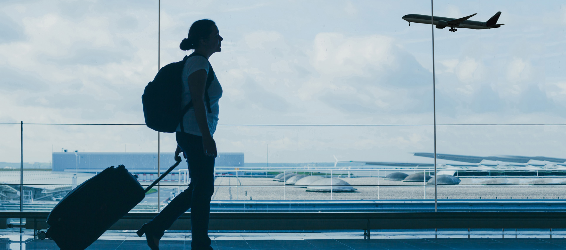Silhouetted traveler with a backpack pulling a suitcase through an airport terminal as a passenger plane approaches outside.