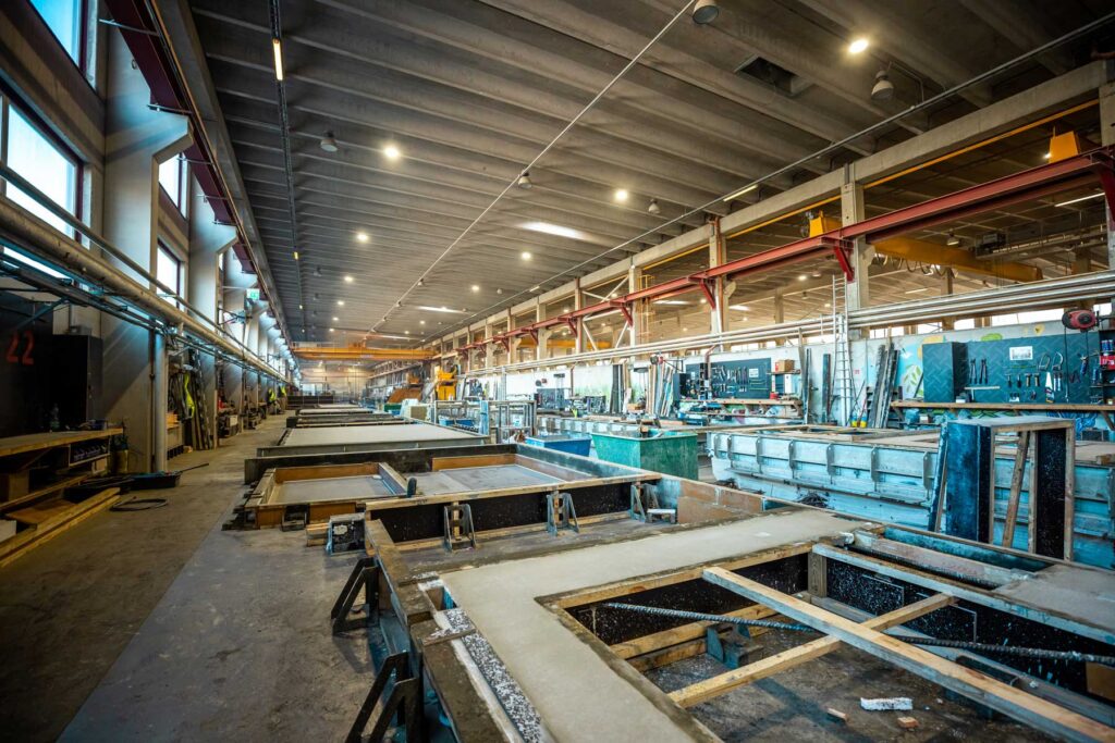 Interior of a large prefabrication facility with concrete molds, workstations, tools, and overhead cranes.