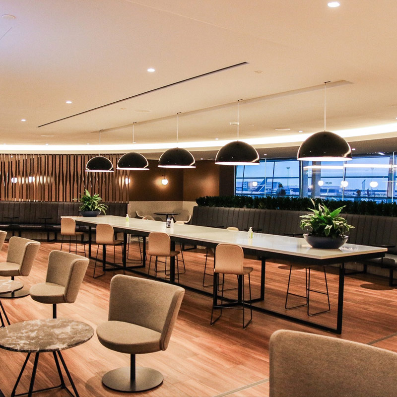 Modern airport lounge with communal tables, upholstered seating, pendant lights, wood flooring, and indoor plants.