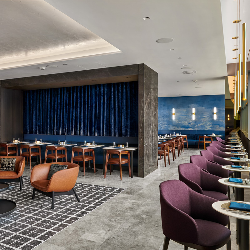 Upscale airport lounge dining area with purple and brown seating, set tables, blue accent walls, and contemporary lighting.