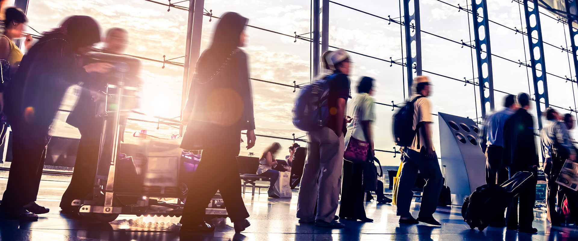 Travelers with luggage move through a sunlit airport terminal with floor-to-ceiling windows.