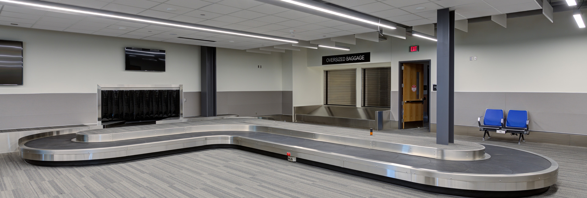 Baggage claim area at Provo Airport featuring a large carousel, oversized baggage pickup, digital screens, and blue seating.