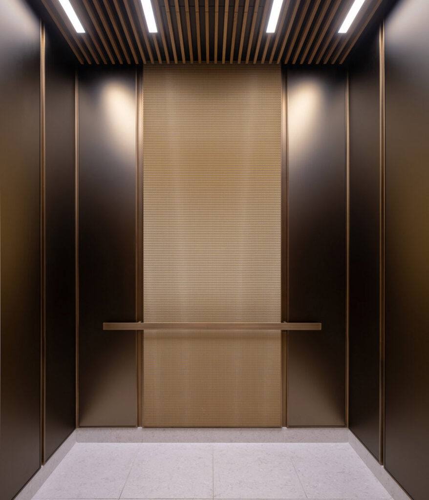 Elevator interior with dark metal wall panels, a textured bronze-toned center panel, horizontal handrail, wood-slat ceiling, linear lights, and light stone flooring.