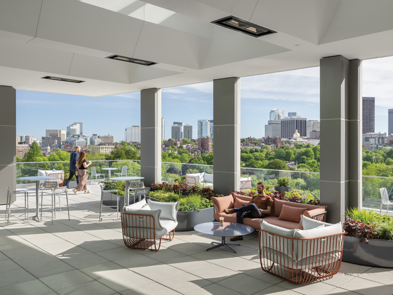 Covered rooftop terrace with lounge seating, dining tables, planted borders, three people, and views of a city skyline.
