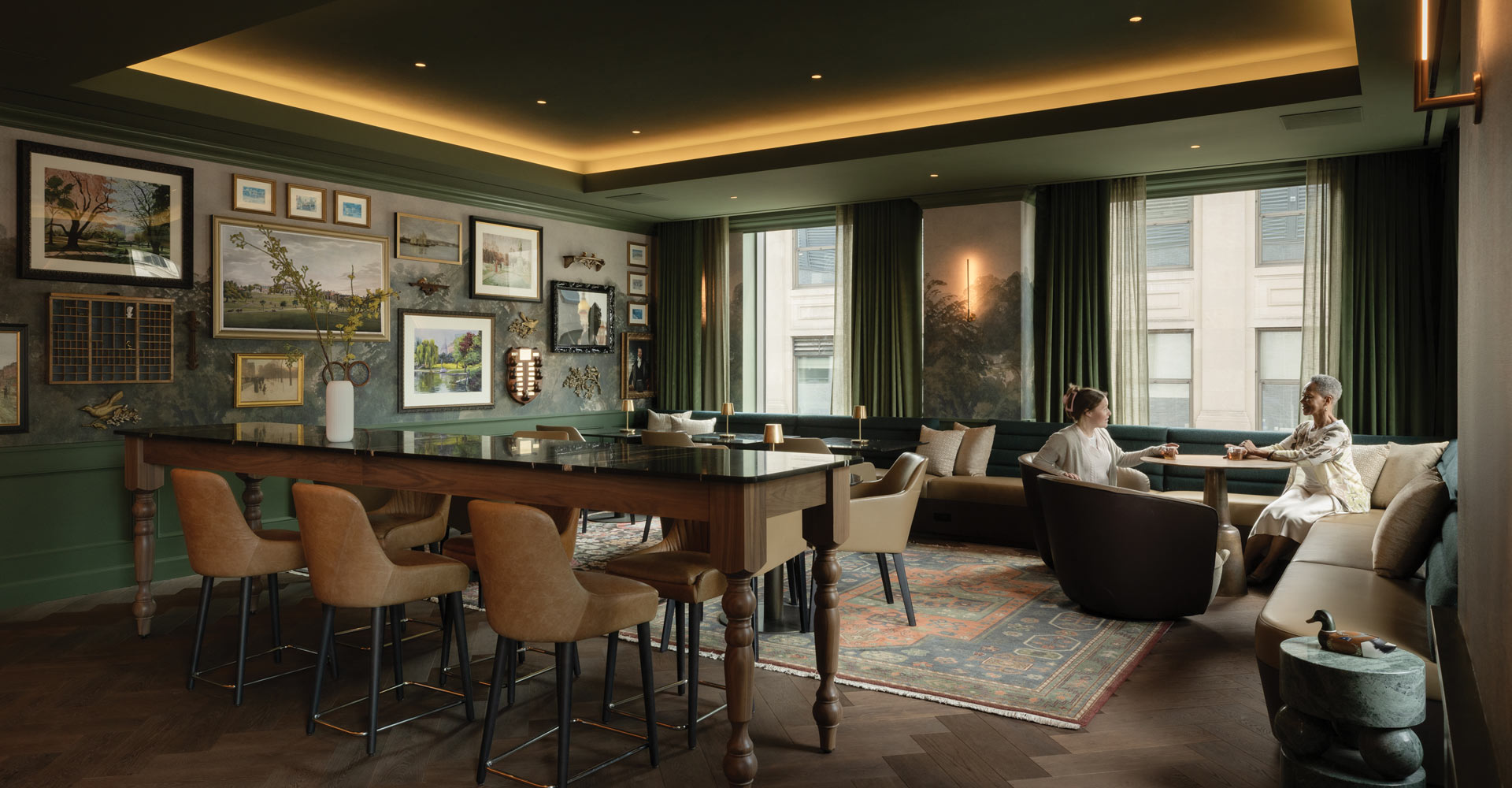 Two people seated at a small table in a lounge with dark green banquette seating, framed artwork, and a long table.