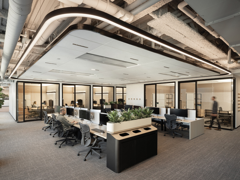 Open-plan office with rows of workstations, indoor planters, glass-walled rooms, exposed ceiling infrastructure, and a suspended rectangular light fixture.