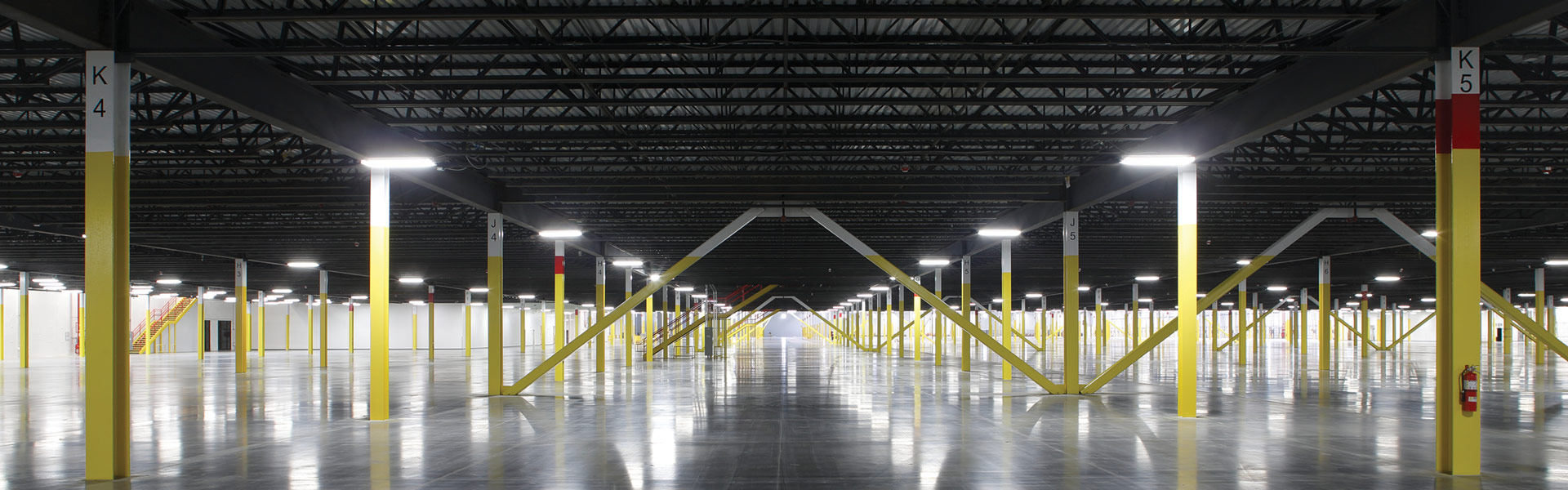 Expansive industrial interior with polished concrete floors, rows of yellow columns, overhead steel framing, and diagonal structural braces.