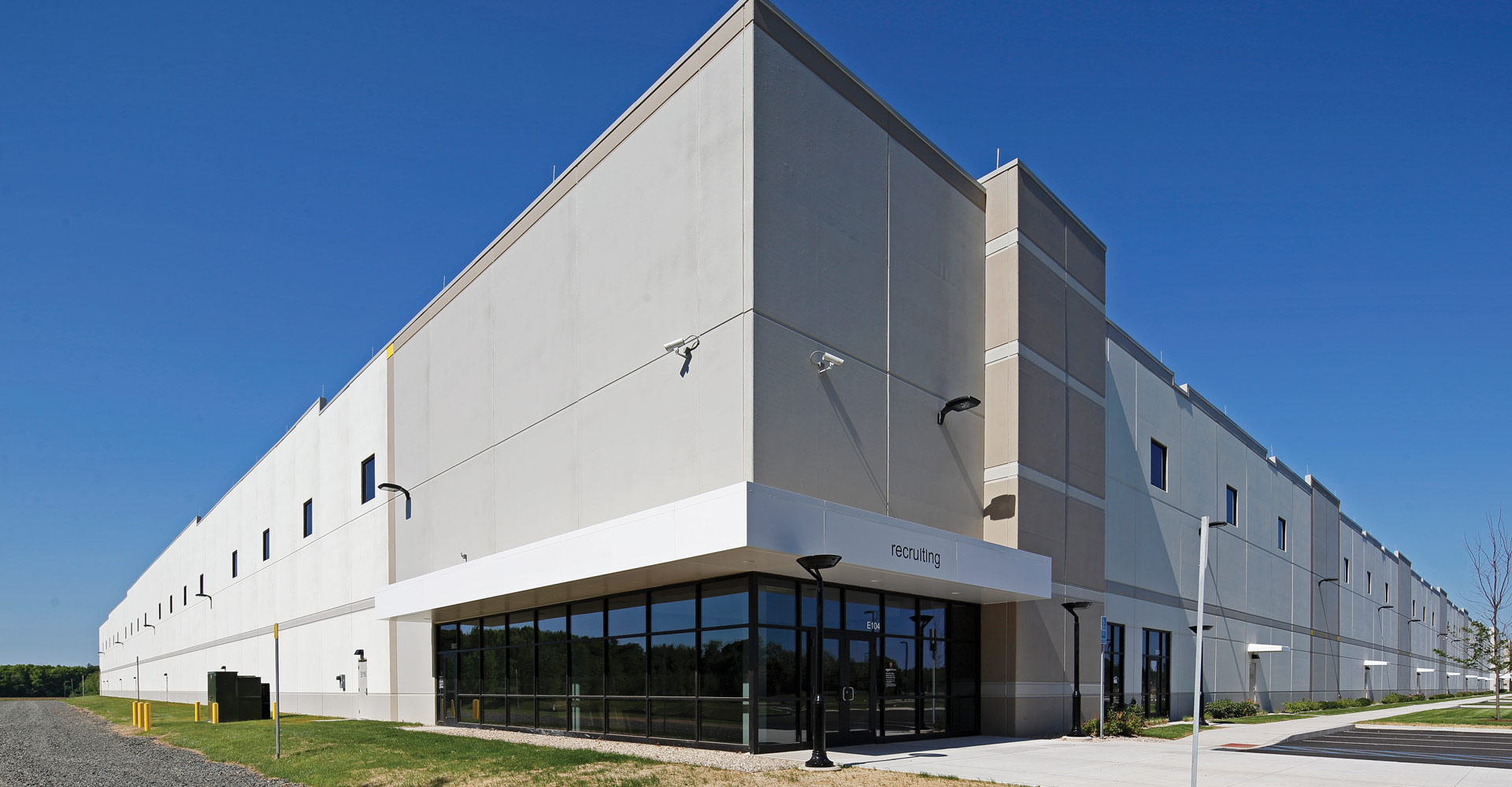Exterior of a large gray industrial building with a glass-fronted entrance and “recruiting” signage.