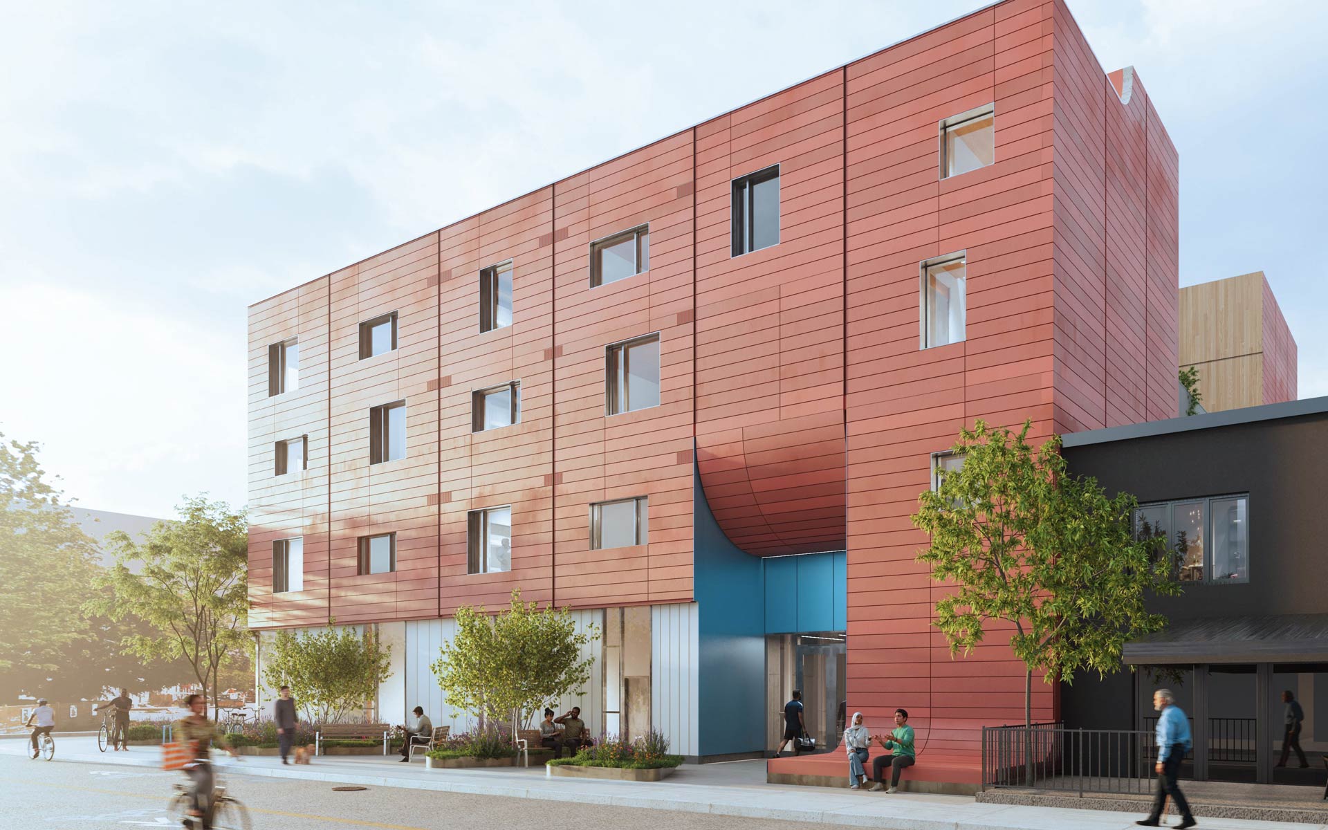 Red-paneled multistory building with a recessed blue entrance, street trees, pedestrians, and cyclists along the sidewalk.
