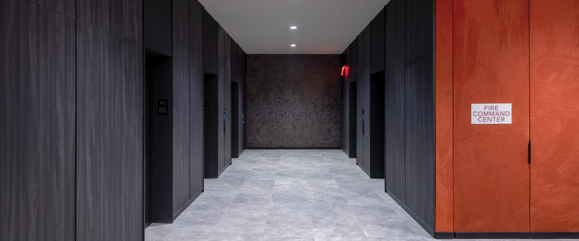 Modern elevator lobby with dark wood-paneled walls, gray tile flooring, and an orange door labeled “Fire Command Center.”