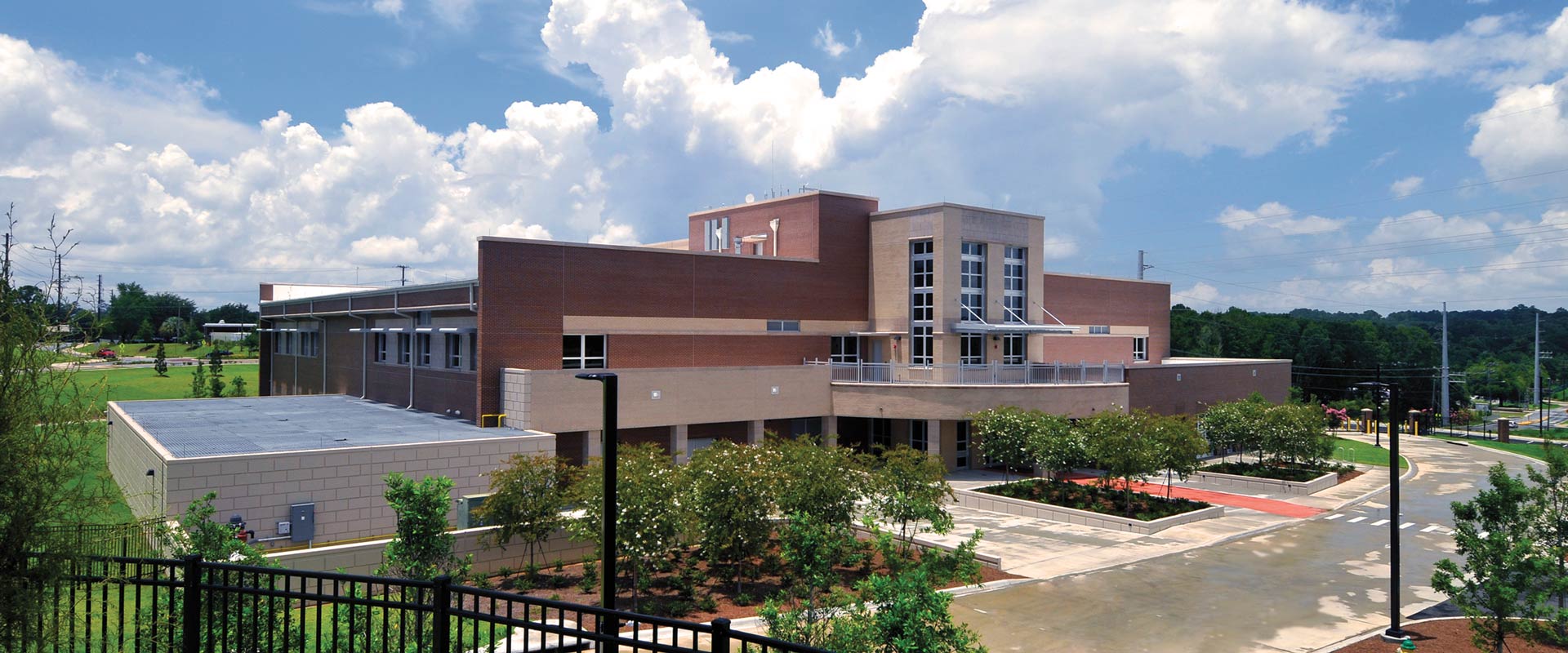 Multistory brick building with a central glass entrance, landscaped grounds, and a paved driveway under a cloudy blue sky.