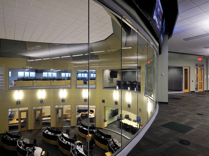 Glass-walled interior overlooking a two-story operations room with curved computer workstations, offices, and conference rooms.