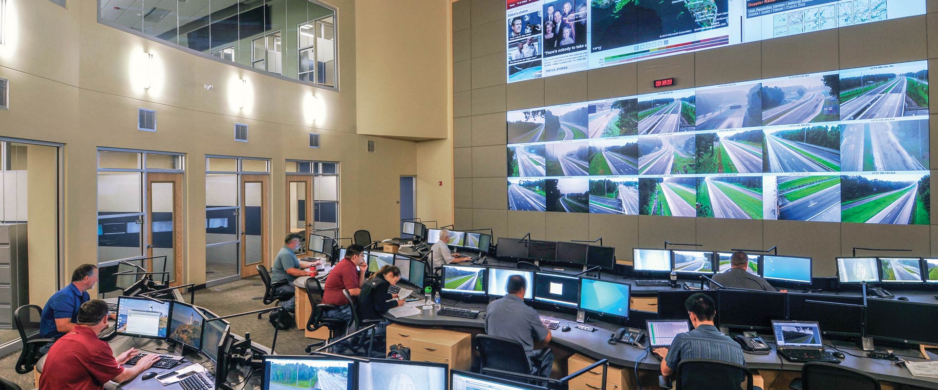 People seated at computer workstations in a control room, facing large wall displays showing roadway camera feeds and weather maps.