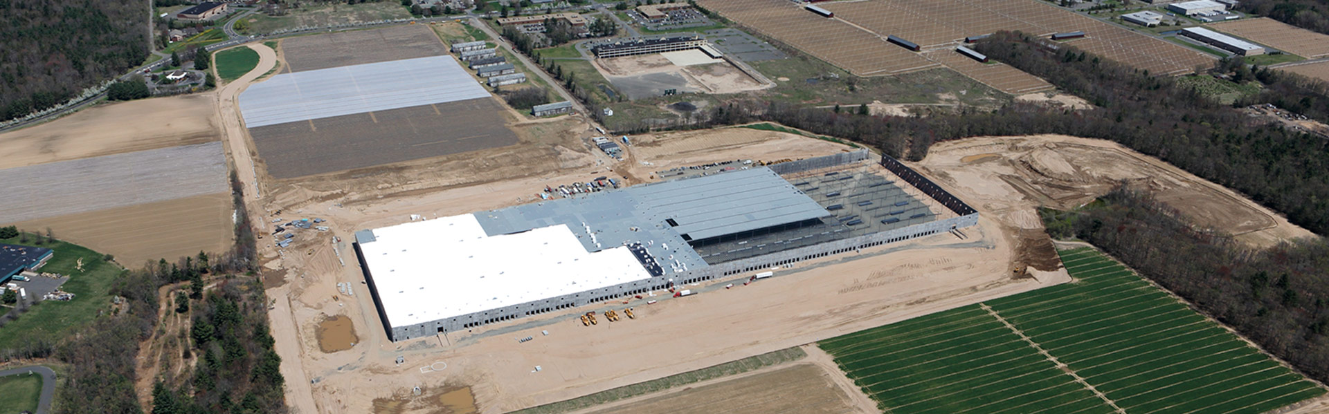 Aerial view of a large building under construction, surrounded by graded earth, fields, wooded areas, and nearby roads.