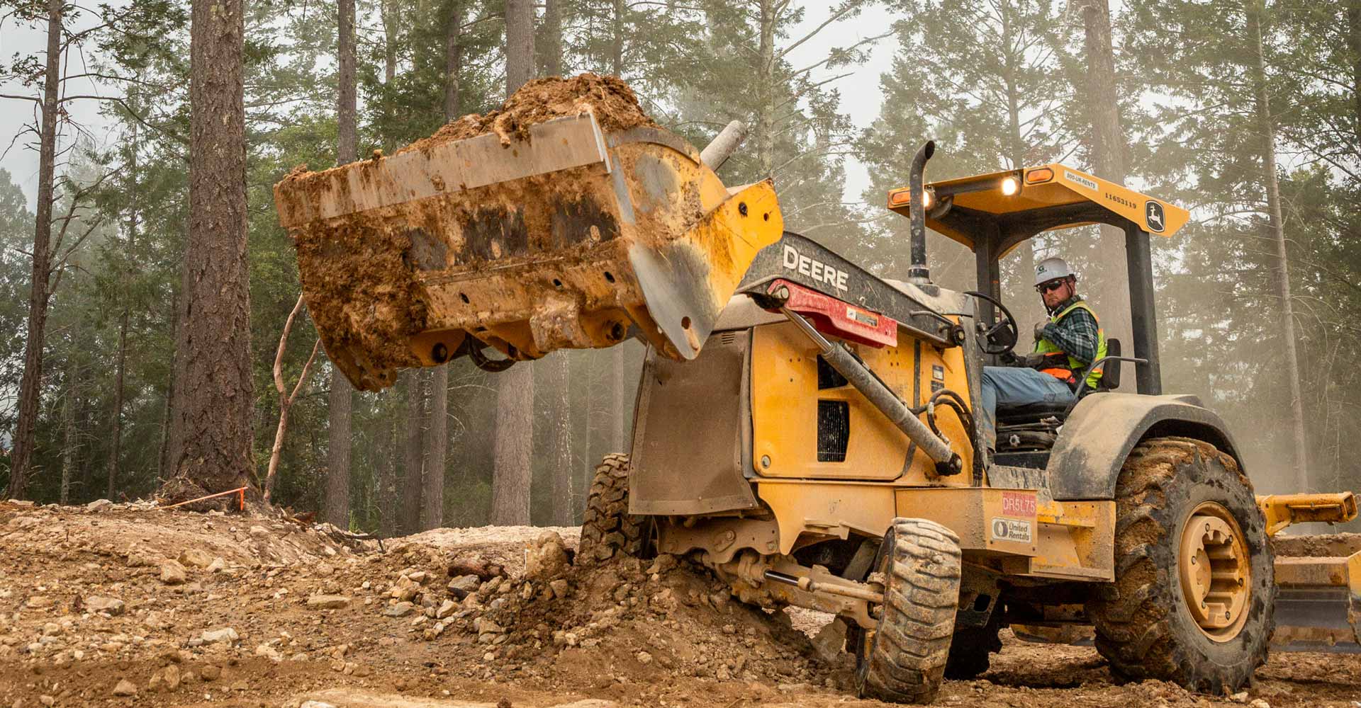 Worker in a hard hat and high-visibility vest operates a yellow John Deere backhoe with a raised bucket of soil at a wooded construction site.