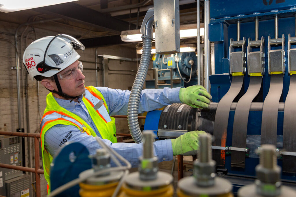 Worker wearing a hard hat, safety glasses, high-visibility vest, and gloves adjusting equipment on a large blue industrial machine.