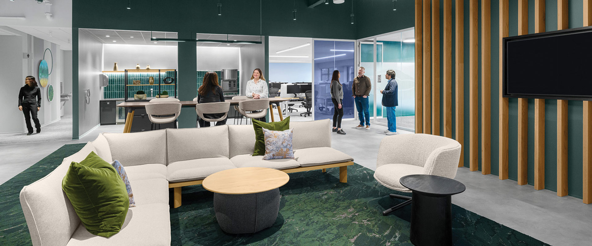 Office lounge with sectional seating, communal tables, a kitchenette, glass-walled work areas, and several people.
