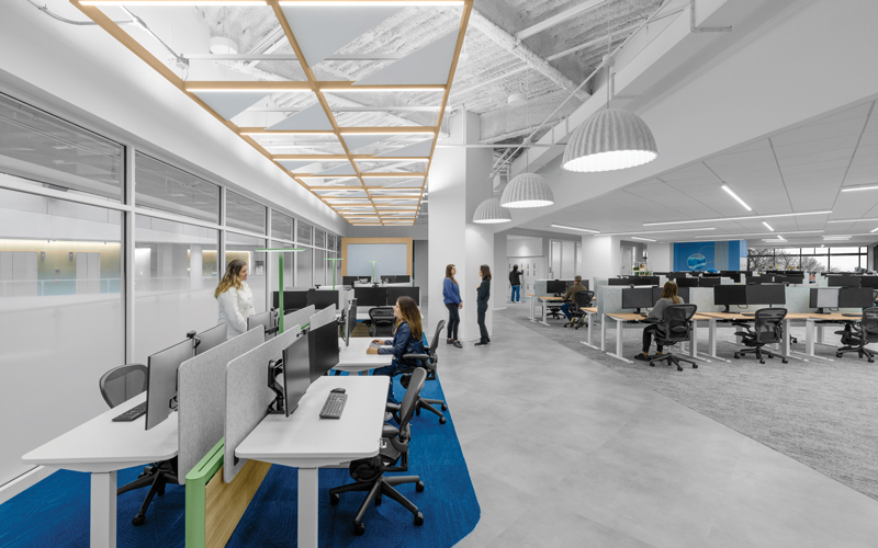 Open office with rows of computer workstations, glass-walled rooms, blue carpet accents, overhead lighting, and several people.