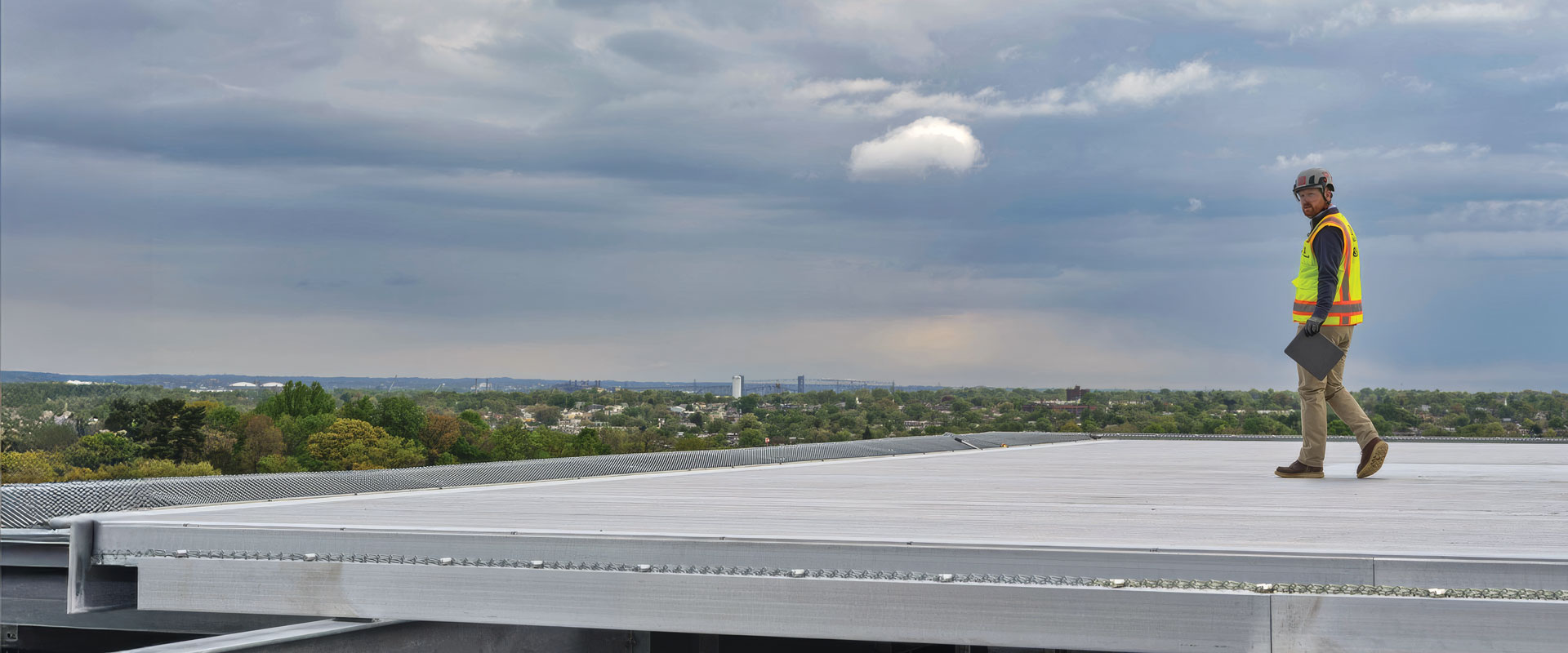 Worker in a high-visibility safety vest and hard hat walking across a large metal rooftop beneath a cloudy sky.
