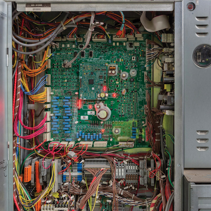 Open electrical control cabinet containing a large green circuit board, bundled multicolored wiring, terminal blocks, and illuminated red indicator lights.