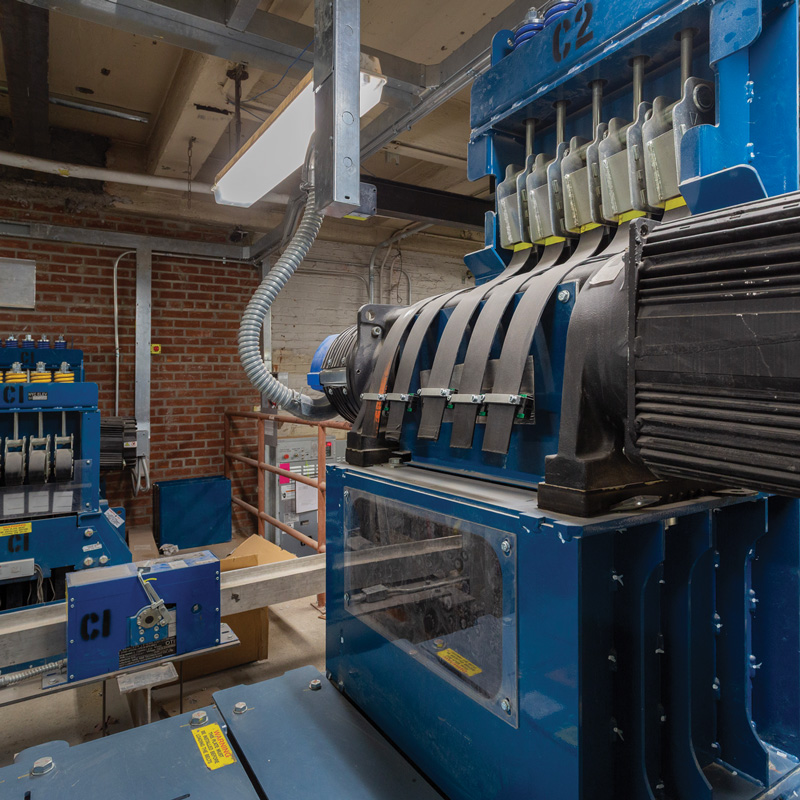 Blue industrial machinery with a large electric motor, multiple drive belts, and metal components in a brick-walled equipment room.