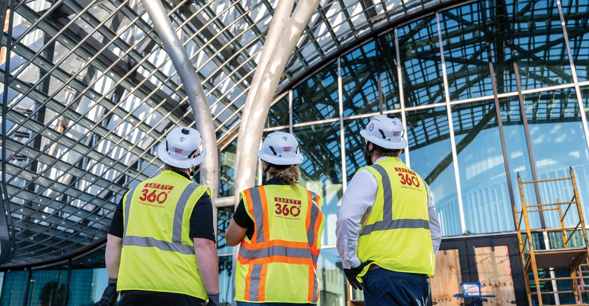 Three people in hard hats and high-visibility “Safety 360” vests stand beneath a curved glass-and-steel building facade.