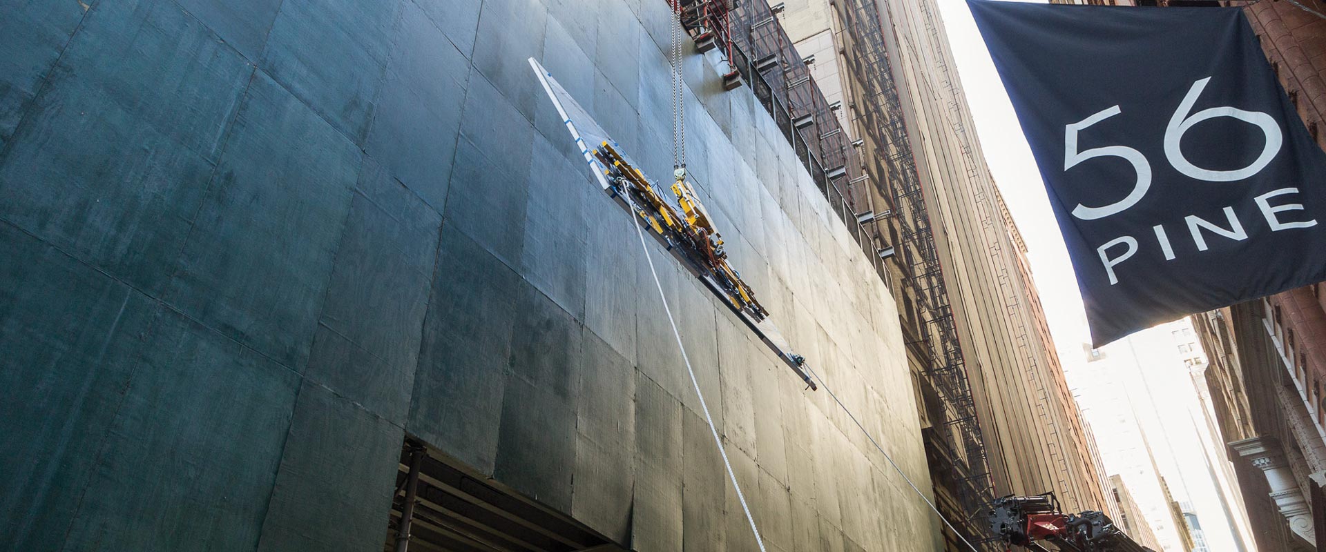 Suspended metal equipment being hoisted beside a covered building facade, with a “56 Pine” flag in the foreground.
