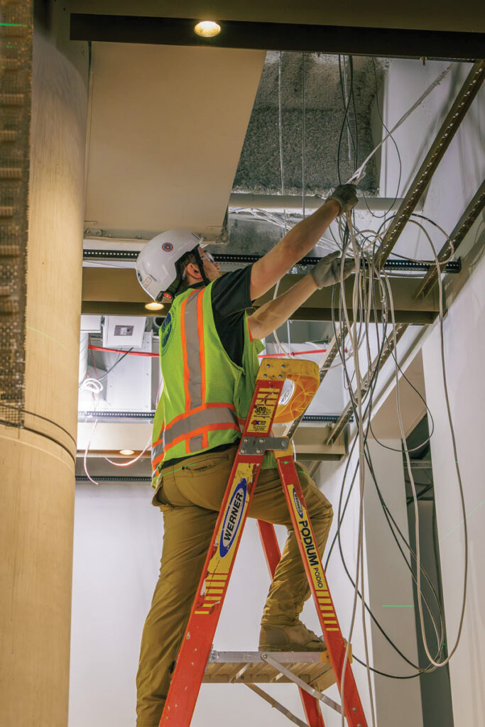 Worker wearing a hard hat and high-visibility vest stands on a red stepladder while arranging cables above an unfinished ceiling.
