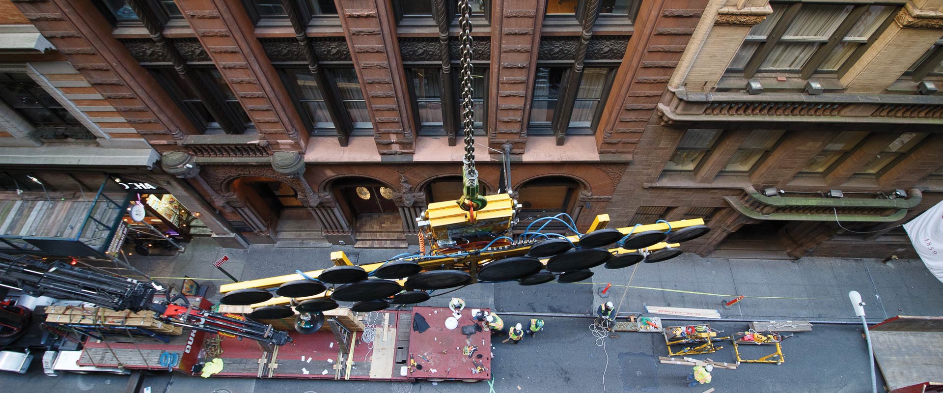 Large suction-cup lifting frame suspended above a closed street, with construction workers and equipment below between masonry buildings.