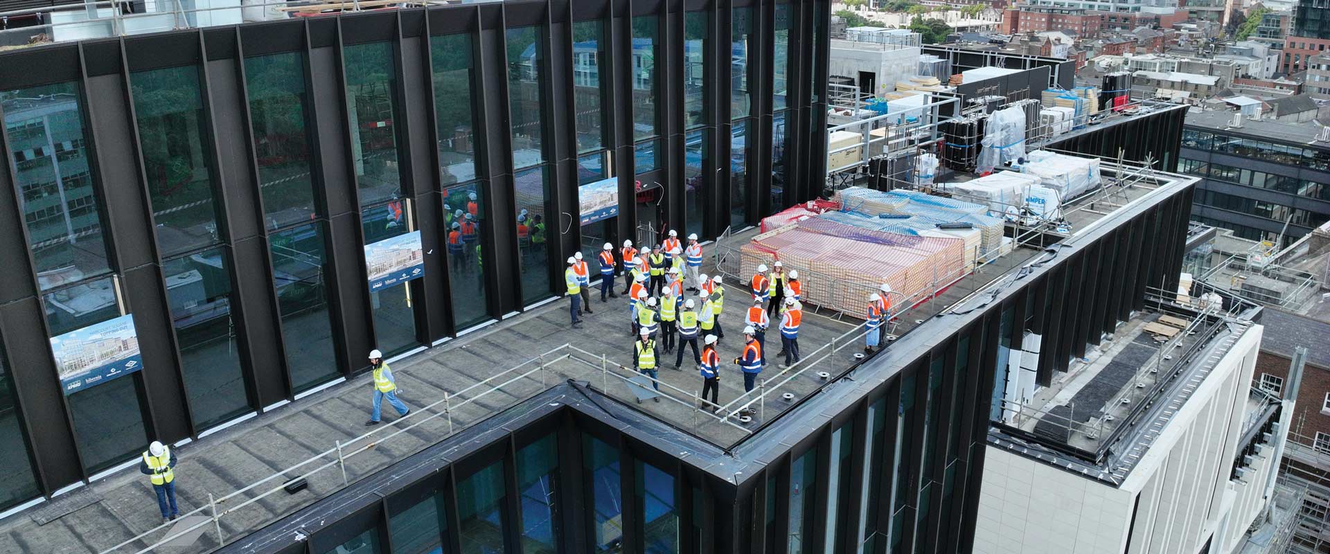 Group of people wearing hard hats and high-visibility vests gathered on the rooftop of a multistory building under construction.