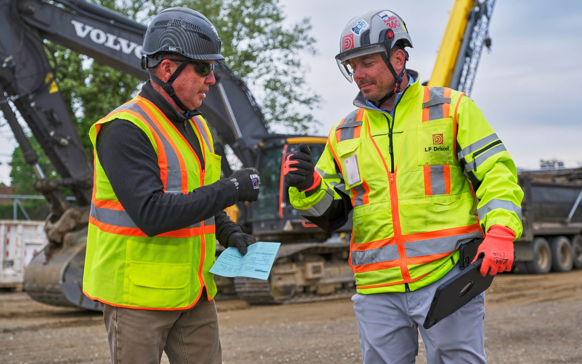 Two construction workers in high-visibility gear review a form beside a Volvo excavator and other heavy equipment at an LF Driscoll jobsite.